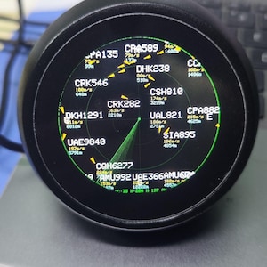 Pode incluir: Um ecrã digital circular preto que mostra uma interface tipo radar com texto e gráficos a verde. O ecrã apresenta vários códigos de voo como CRK546, DHK238 e UAL821, juntamente com dados numéricos de velocidade em m/s e altitude em metros.