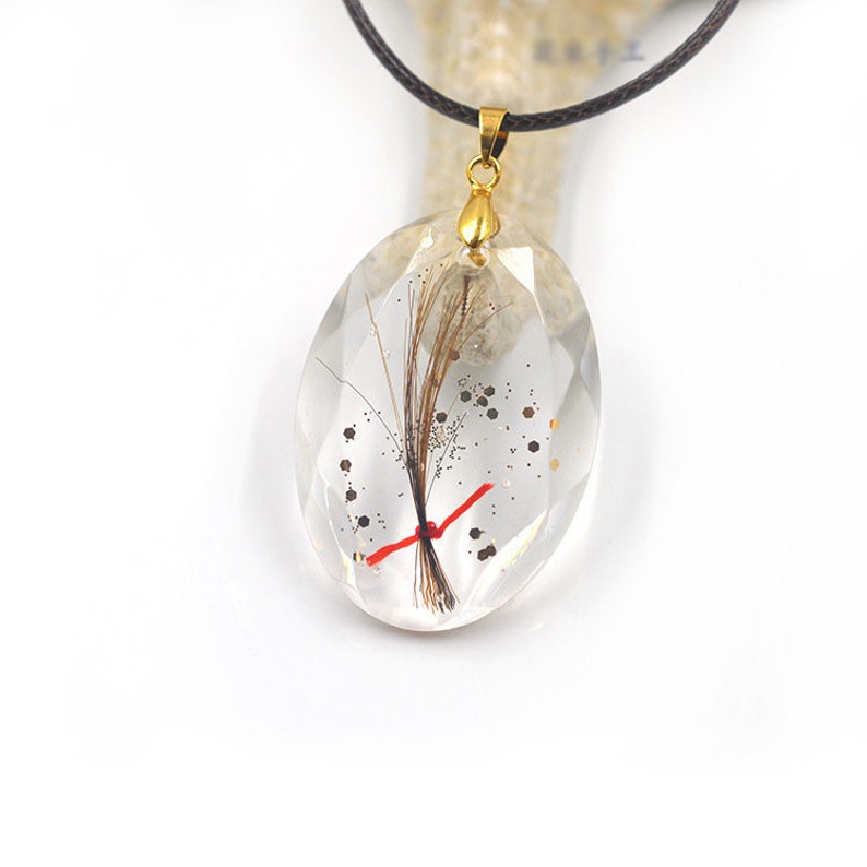 May include: A gold chain necklace with a clear oval pendant containing a small amount of hair, red thread, and gold glitter.
