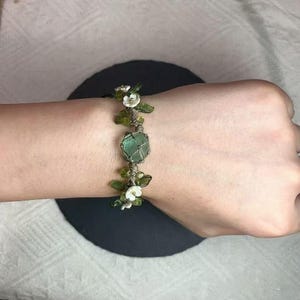 May include: A green gemstone bracelet with white flower accents and a brown cord.