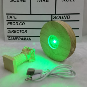 Könnte beinhalten: Ein Holzsockel mit einem runden Ausschnitt, der ein kleines Objekt mit grünem Licht beleuchtet. Der Sockel ist mit einem weißen USB-Kabel verbunden.