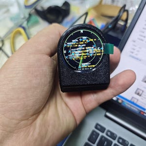 Puede incluir: Un dispositivo electrónico negro de forma semicircular con una pantalla digital circular. La pantalla muestra una interfaz tipo radar con varios datos de vuelo, incluyendo etiquetas como N9379L, 3300 ft y AIR 29 N111 sobre un fondo oscuro.