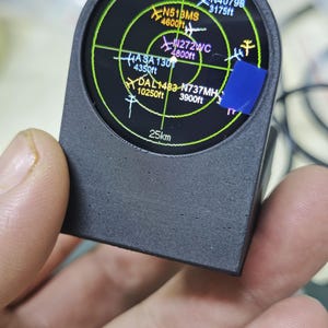 May include: A black, 3D-printed, handheld radar display device featuring a circular screen with a green grid overlay. The screen displays various aircraft identifiers like N40798, N513MS, N272WC, ASA130, DAL1483, and N737MH, along with altitude data and a 25km scale.