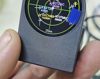 May include: A black, 3D-printed, handheld radar display device featuring a circular screen with a green grid overlay. The screen displays various aircraft identifiers like N40798, N513MS, N272WC, ASA130, DAL1483, and N737MH, along with altitude data and a 25km scale.