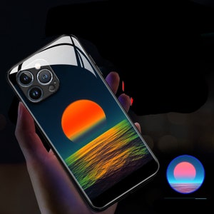 Puede incluir: Una funda negra para iPhone con una escena de puesta de sol y océano. La funda tiene una parte posterior transparente y un marco negro. La puesta de sol es naranja y amarilla, y el océano es azul y verde.