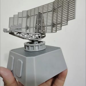 Könnte beinhalten: Ein graues Kunststoffmodell einer Radarantenne mit einem großen, rechteckigen Gitterarray, das auf einer drehbaren Stützstruktur montiert ist. Die Baugruppe sitzt auf einer massiven, trapezförmigen Basis mit zwei rechteckigen Paneelen.