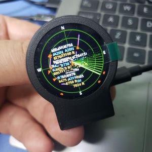 Könnte beinhalten: Ein rundes schwarzes elektronisches Gerät mit einem kreisförmigen Bildschirm, der eine grüne Radarschnittstelle anzeigt. Der Bildschirm zeigt verschiedene alphanumerische Codes, Höhendaten in Fuß und Richtungskennzeichnungen. Eine kleine grüne Lasche ist an der rechten Seite befestigt.
