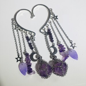 May include: A pair of silver ear cuffs with dangling charms. The charms include amethyst stones, crescent moons, stars, and leaves. The amethyst stones are in a cage-like design.