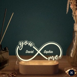 Puede incluir: Símbolo de infinito blanco con las palabras "you" y "me" a cada lado. Los nombres "David" y "Sophia" están escritos dentro del símbolo. El símbolo de infinito está iluminado con luz blanca y se asienta sobre una base de madera.