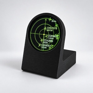 Puede incluir: Un soporte negro en forma de L con una pantalla digital circular que imita un radar de aviación. La pantalla muestra trayectorias de vuelo en verde brillante, iconos de aviones y etiquetas de texto como CHH7078, CQH8631, CCA2869 y CSH9357.