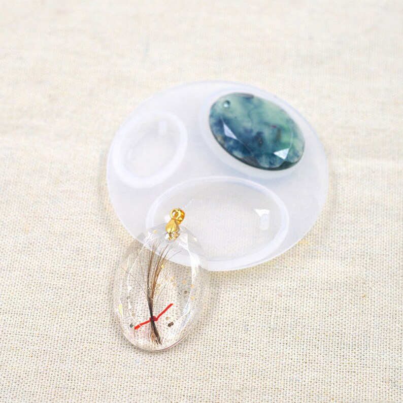 May include: A clear oval resin pendant with a gold bail and a small red string and brown hair inside. The pendant is sitting on a white silicone mould with two other oval moulds and a round mould. The mould has a green and blue stone in one of the oval moulds.