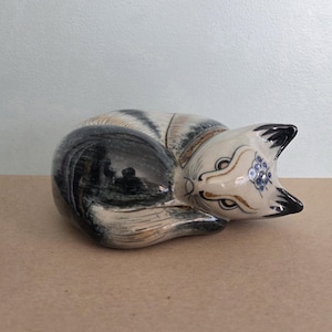 May include: A ceramic cat figurine in a curled-up position. The cat has a cream-colored body with black and gray stripes and accents. The face features black ears, brown and blue decorative details, and closed eyes. The figurine is on a brown surface.