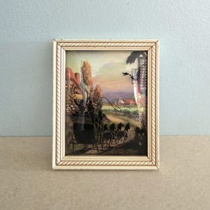 May include: Framed vintage silhouette art depicting a horse-drawn carriage scene. The artwork features a cream-colored frame with a rope-like border. The background shows a colorful landscape with a house and trees.