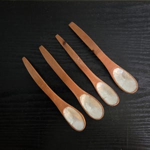 May include: A set of four wooden spoons with elongated handles and oval-shaped bowls. Each spoon features a smooth, light brown wooden handle and a distinct, iridescent white mother-of-pearl inlay lining the interior of the bowl section.