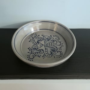 May include: A round, ceramic serving dish with a light gray base and a dark blue floral design in the center. The rim and the design are outlined in dark blue. The dish is approximately 10 inches in diameter.