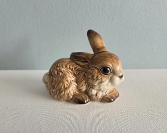May include: A small ceramic figurine of a crouching rabbit with textured brown fur, dark eyes, and one upright ear. The rabbit has a cream-colored chest and muzzle, with detailed shading throughout its body, capturing a realistic posture in a seated position.