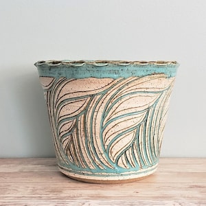 May include: A ceramic planter with a light blue rim and a white and light blue leaf pattern. The planter is sitting on a light wood surface.