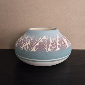May include: A small, round, ceramic vase with a light blue exterior. The vase features a band of white and purple geometric designs around the top. A white stripe encircles the base. The interior is white. The vase sits on a black surface.