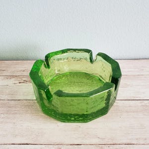 May include: A vintage green glass ashtray with a textured surface. The ashtray has a unique, irregular shape with a series of notches around the rim. The color is a vibrant, translucent green, and the glass appears thick and durable.
