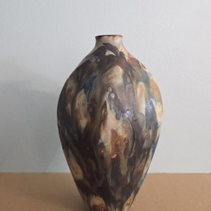 May include: A handcrafted ceramic vase with a bulbous body and a narrow neck. The vase features a unique glaze with a blend of brown, beige, blue, and green hues, creating a marbled effect. The vase is approximately 10 inches tall.