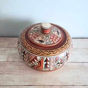 May include: A round, ceramic jar with a lid, decorated with geometric patterns in black, white, and red on a brown background. The lid has a small, white knob.