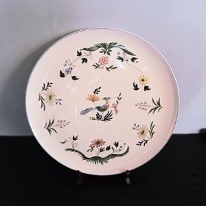 May include: A round, cream-colored decorative plate with a silver rim. The plate features a central illustration of a bird and floral designs in shades of green, yellow, pink, and blue. The plate is displayed on a black stand.