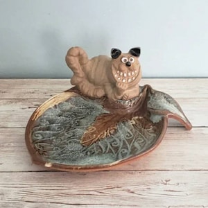 May include: A ceramic dish shaped like a leaf with a whimsical cat figurine on top. The cat has a wide grin and black eyes. The dish has a blue and brown glaze with a swirling pattern.