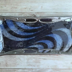 May include: Rectangular decorative tray with a silver-colored metal frame. The tray's interior features a blue and black swirling pattern. The tray is placed on a light-colored wooden surface.