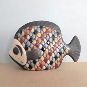May include: A ceramic fish figurine featuring a matte dark gray head, tail, and fins. The body is covered in a mosaic-like pattern of small, rounded scales in shades of red, yellow, blue, and cream. The face has a light cream-colored area.