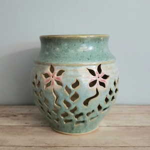 May include: A teal blue ceramic candle holder with a delicate floral design. The holder has cut-out flowers and leaves, creating a light and airy feel.