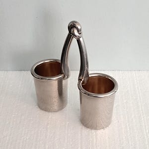 May include: A silver-colored double container with a handle. The handle is a curved design that connects the two cylindrical cups at the top with a knot. The interior of the cups has a copper-colored finish. The object is on a white surface.