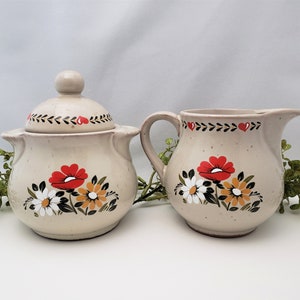 May include: A white ceramic sugar bowl and creamer set with a floral design. The design features red, white, and yellow flowers with green leaves. The set has a vintage look.