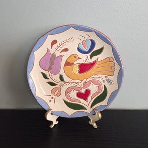 May include: A round ceramic plate with a scalloped blue rim and a cream-colored center. The design features a central yellow bird with red wing accents, surrounded by stylized floral motifs in purple, pink, and green, and a red heart shape.