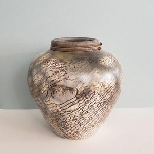 May include: A decorative vase with a textured, metallic finish. The vase has a bulbous body and a narrow neck with a bronze-colored rim. The surface features a pattern of intersecting lines and mottled brown and silver tones.