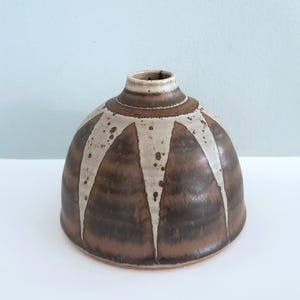 May include: A handcrafted ceramic vase with a rounded base and a narrow neck. The vase features a brown and white geometric pattern, with white triangular shapes radiating from the top. The vase is approximately 6 inches tall.