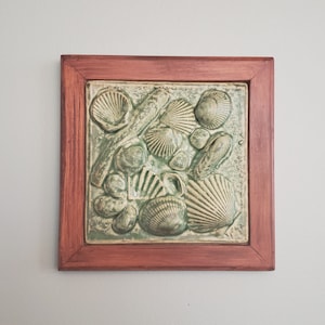 May include: A framed ceramic tile with a green and white seashell design. The tile is surrounded by a dark wood frame.