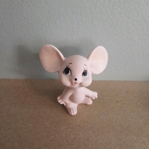 May include: A light pink ceramic mouse figurine with large ears, black eyes, and delicate eyelashes. The mouse is in a seated position with its arms outstretched. The figurine has a smooth finish and is likely a decorative item.