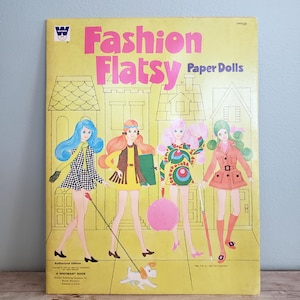 May include: Vintage "Fashion Flatsy Paper Dolls" book with four illustrated paper dolls in colorful outfits. The cover is yellow with pink text and a small white dog illustration. The book is an authorized edition.