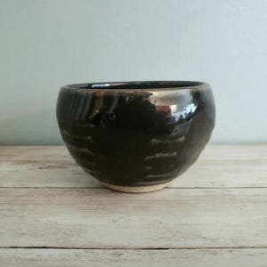May include: A small, black ceramic bowl with a slightly textured surface. The bowl has a light brown rim and a glossy finish.
