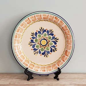 May include: A white ceramic plate with a blue, green, and yellow floral design. The plate has a scalloped edge and is decorated with a peach-colored border.