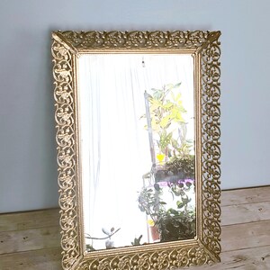 May include: A gold-toned ornate mirror with a decorative frame. The mirror is rectangular and has a reflective surface. The frame is made of metal and has a floral design.