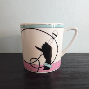 May include: White ceramic mug with a black graphic of a hand holding a microphone. The mug has a light blue and pink band around the top and bottom. The handle is white.