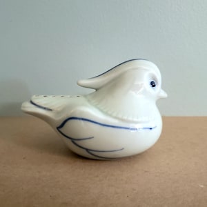 May include: A white ceramic bird figurine with blue accents. The bird has a rounded body, a small beak, and a curved crest. Blue lines detail the wings and tail. The bird has small holes on its back.