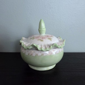 May include: A small, round, ceramic trinket box with a lid. The box is light green with a ruffled edge and a floral design on the lid. The lid has a small, pointed finial. The box sits on a small base.