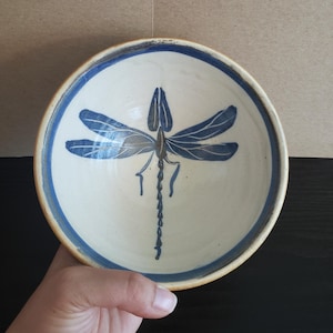 May include: A ceramic bowl with a cream-colored interior and a blue dragonfly design. The rim is outlined in blue. The bowl is round and appears to be handmade.