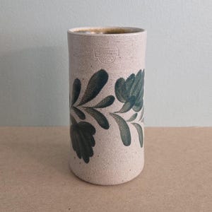 May include: A cylindrical ceramic vase with a speckled, off-white finish. The vase features a dark teal floral design wrapping around the body. The top rim is a lighter shade of tan. The vase has a stamp near the top.