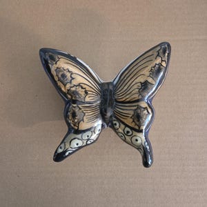 May include: A ceramic butterfly figurine with a glossy finish. The butterfly has black and gold wings with intricate patterns and a black body. The wings are detailed with floral designs and the body has a textured appearance.