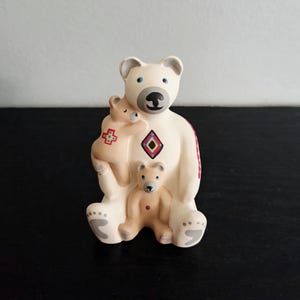 May include: A ceramic figurine of a mother bear with two cubs. The mother bear is white with a black nose and blue eyes. The cubs are tan. The mother bear has a diamond pattern on her chest and a red and white pattern on her side.