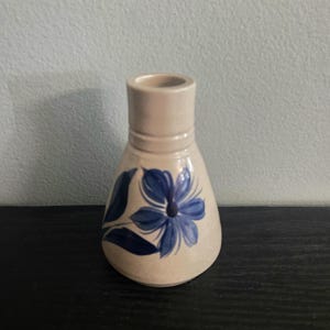 May include: A small, beige ceramic vase with a blue floral design. The vase has a rounded body and a cylindrical neck. The blue flower is painted on the front of the vase, with leaves on the side. The vase is approximately 4 inches tall.