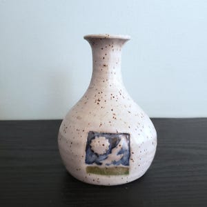 May include: A small, speckled ceramic vase with a rounded body and a narrow neck. The vase is off-white with brown speckles and features a square, blue and green design. The design appears to depict a bird or dove.