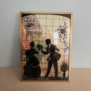 May include: Framed silhouette art depicting a man offering flowers to a woman. The black silhouettes are set against a backdrop of a building and hanging plants. The frame is gold-colored.